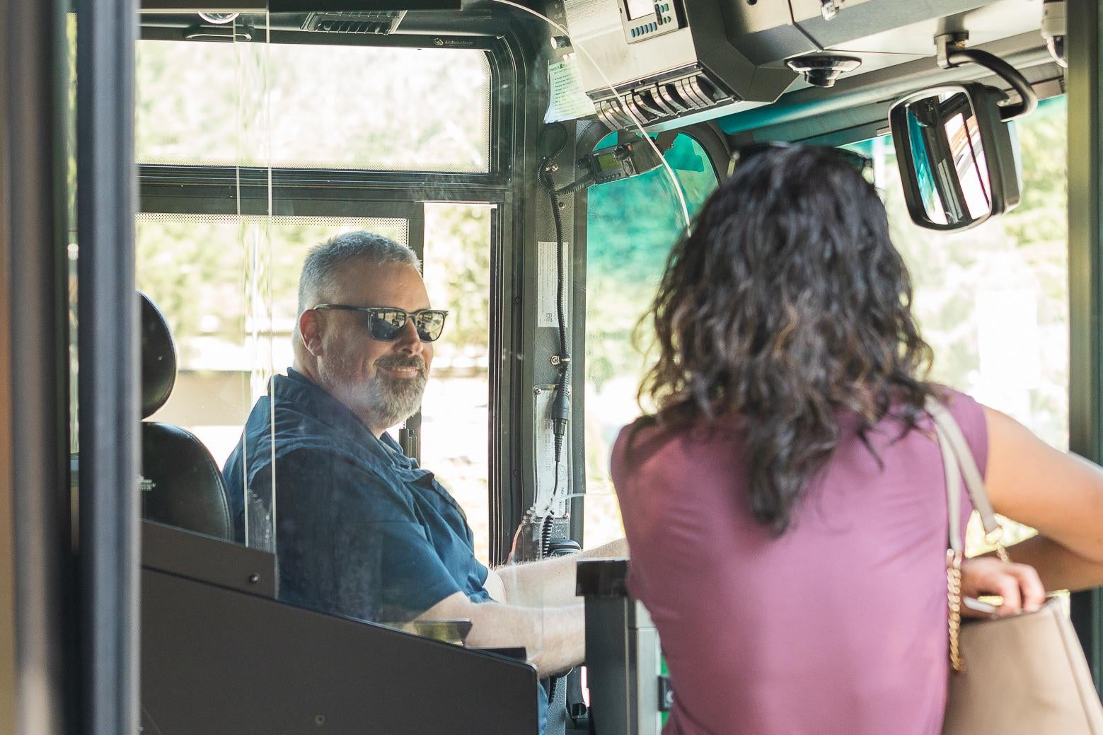 A rider boarding a bus, viewed from behind as they step on board. A driver is smiling at them.