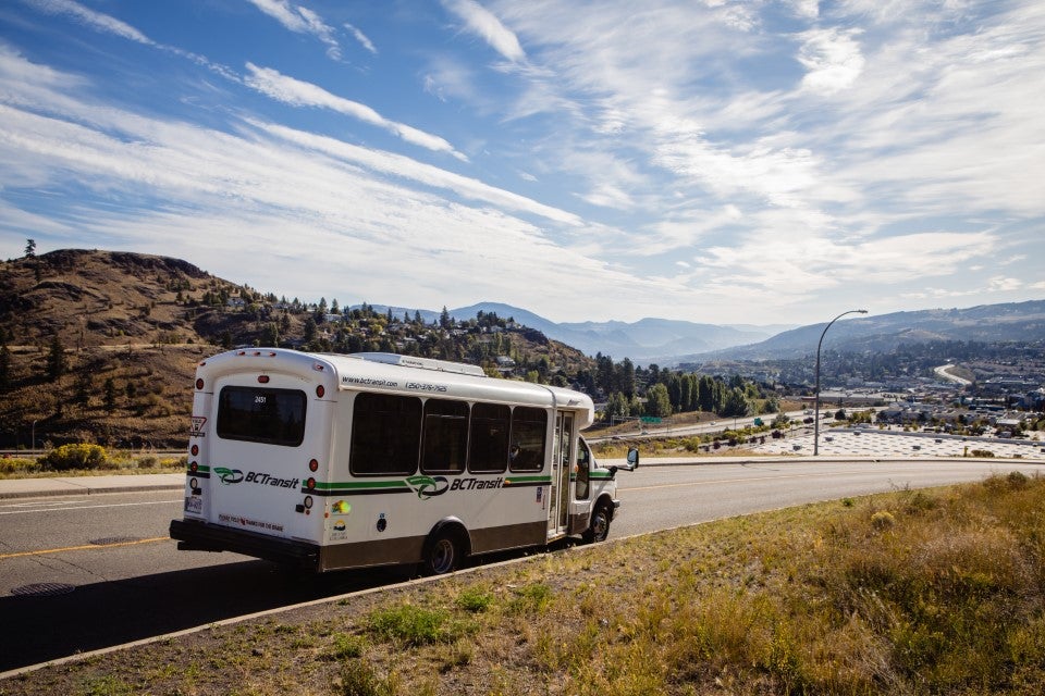 A BC Transit bus drives toward Kamloops, framed by wide skies, hills, and the open Thompson Valley road ahead.