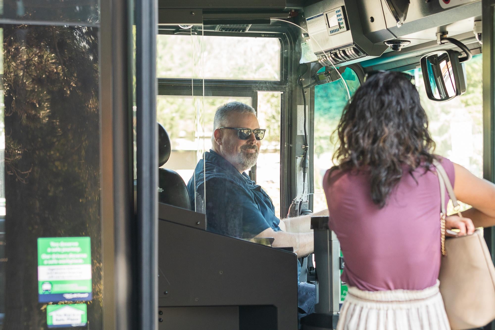 A rider boarding a bus, viewed from behind as they step on board. A driver is smiling at them.