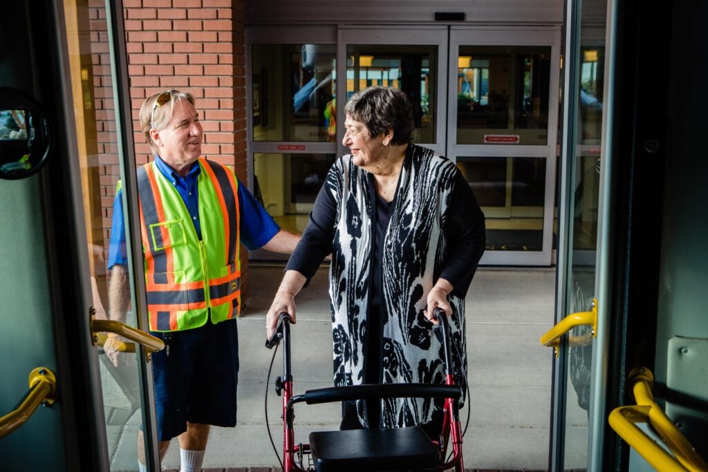 Woman with stroller boards city bus as operator assists at low-floor entrance.