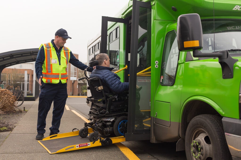 handyDART bus with ramp deployed. Passenger on a wheelchair getting on the bus with the operator helping.