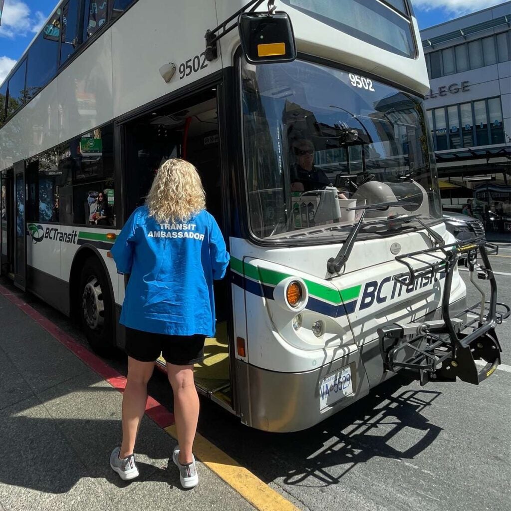 A BC Transit Ambassador wearing a bright blue shirt stands at the open front doors of a double-decker bus, interacting with the driver.