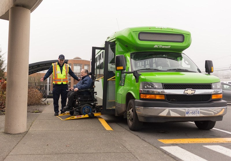 A driver helps a passenger in a wheelchair exit a handyDART bus via a ramp.