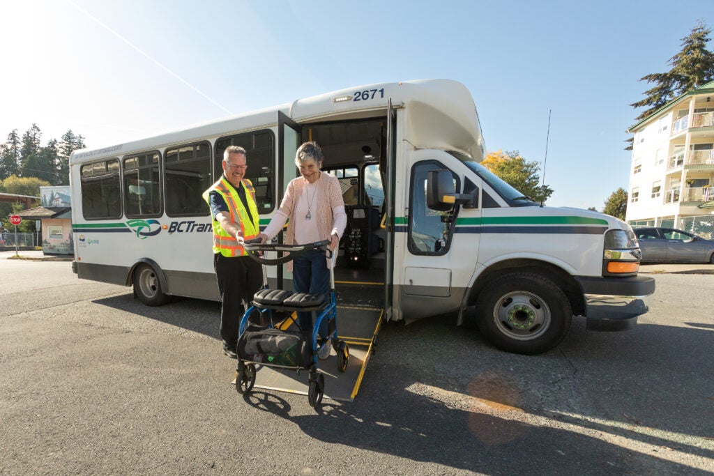 A driver helping a passenger using a mobility aid exit a handyDART bus. The bus has its wheelchair ramp extended.