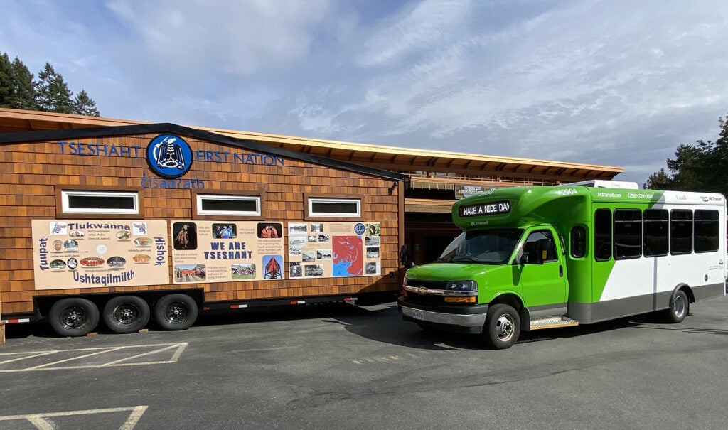 A BC Transit transit bus parked next to a Tsteshaht First Nation 'mini-longhouse' mobile interpretative trailer.