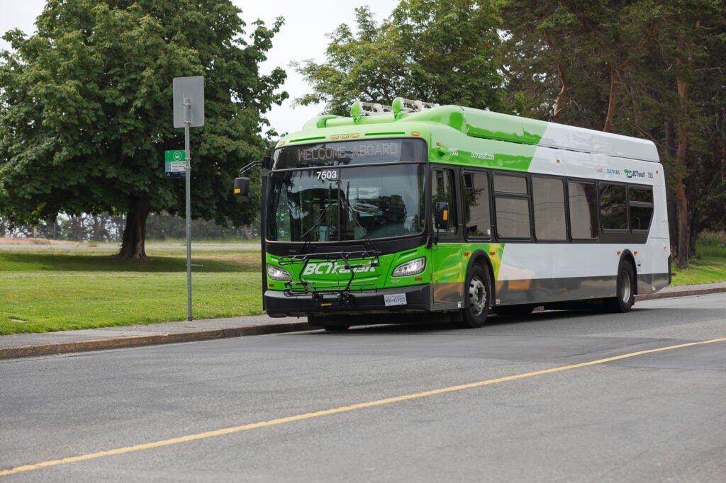 A battery electric bus at a bus stop in front of a park.