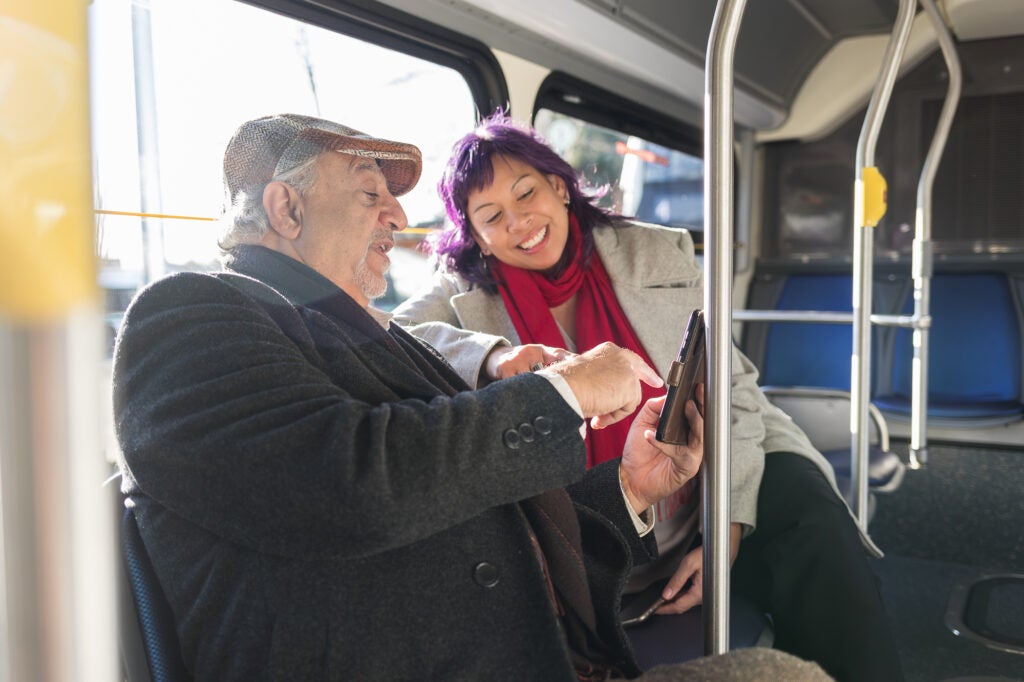 A man and a woman sit on a bus, smiling and looking at a phone together.
