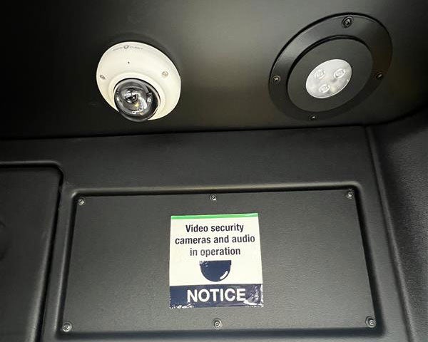 A CCTV camera in the ceiling of a BC Transit bus. The sign below reads "Video security cameras and audio in operation".