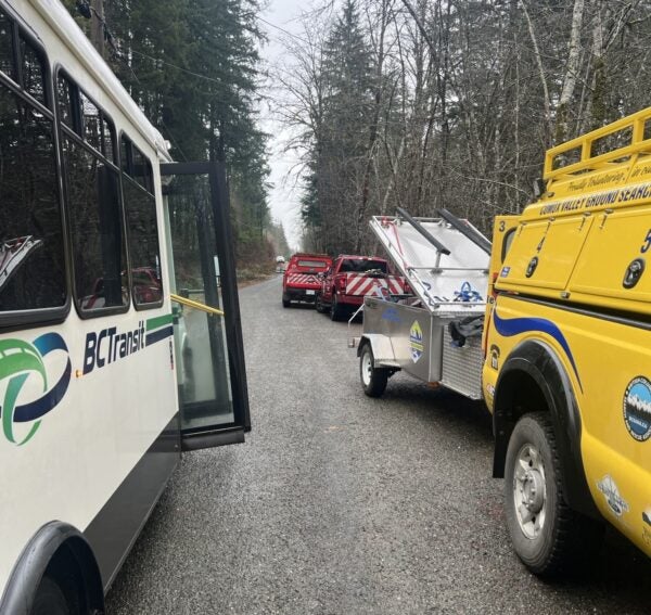 A BC Transit bus and search and rescue vehicles are staged on a rural road in the forest.