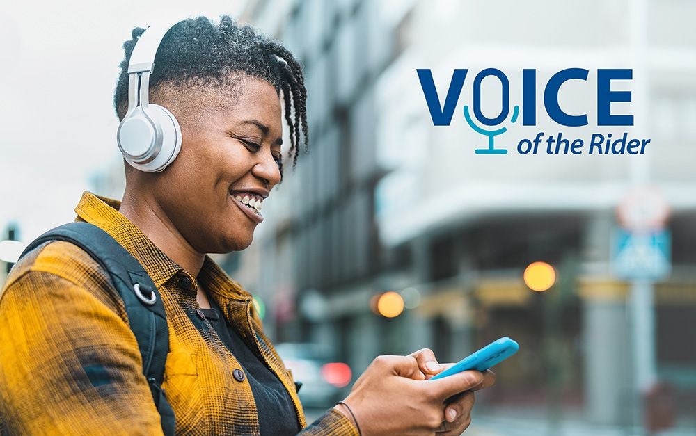 Person wearing headphones and a yellow jacket, smiling while looking at a smartphone on a city street. The Voice of the Rider logo is in the top right corner.