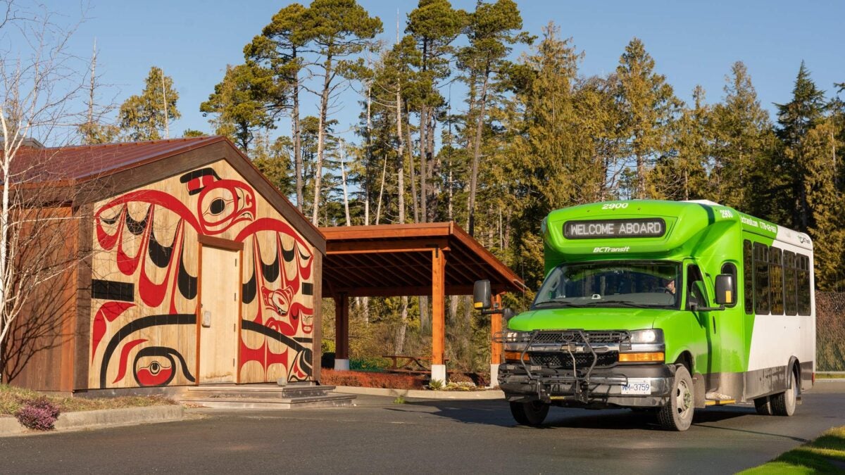 A BC Transit bus is stopped in front of a cedar clad building at the Cixʷatin Centre in Hitacu. Art on the building shows a thunderbird on a whale hunt and also a wolf.