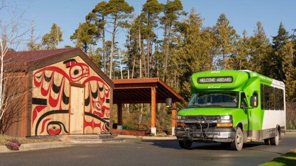 A BC Transit bus is stopped in front of a cedar clad building at the Cixʷatin Centre in Hitacu. Art on the building shows a thunderbird on a whale hunt and also a wolf.