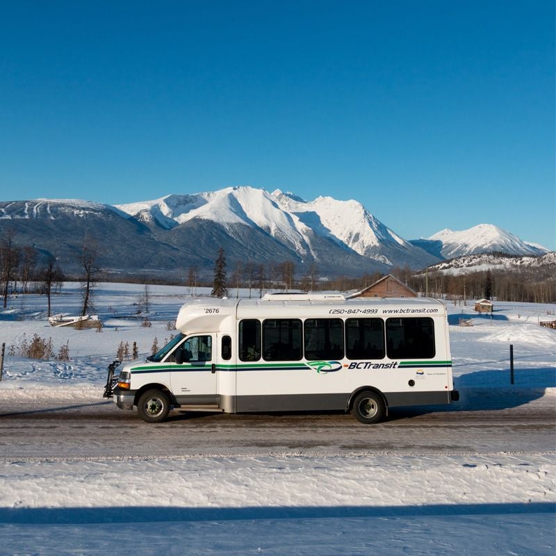 A light duty BC Transit bus on a snowy road in a rural farm area. Snowy mountains are in the background.