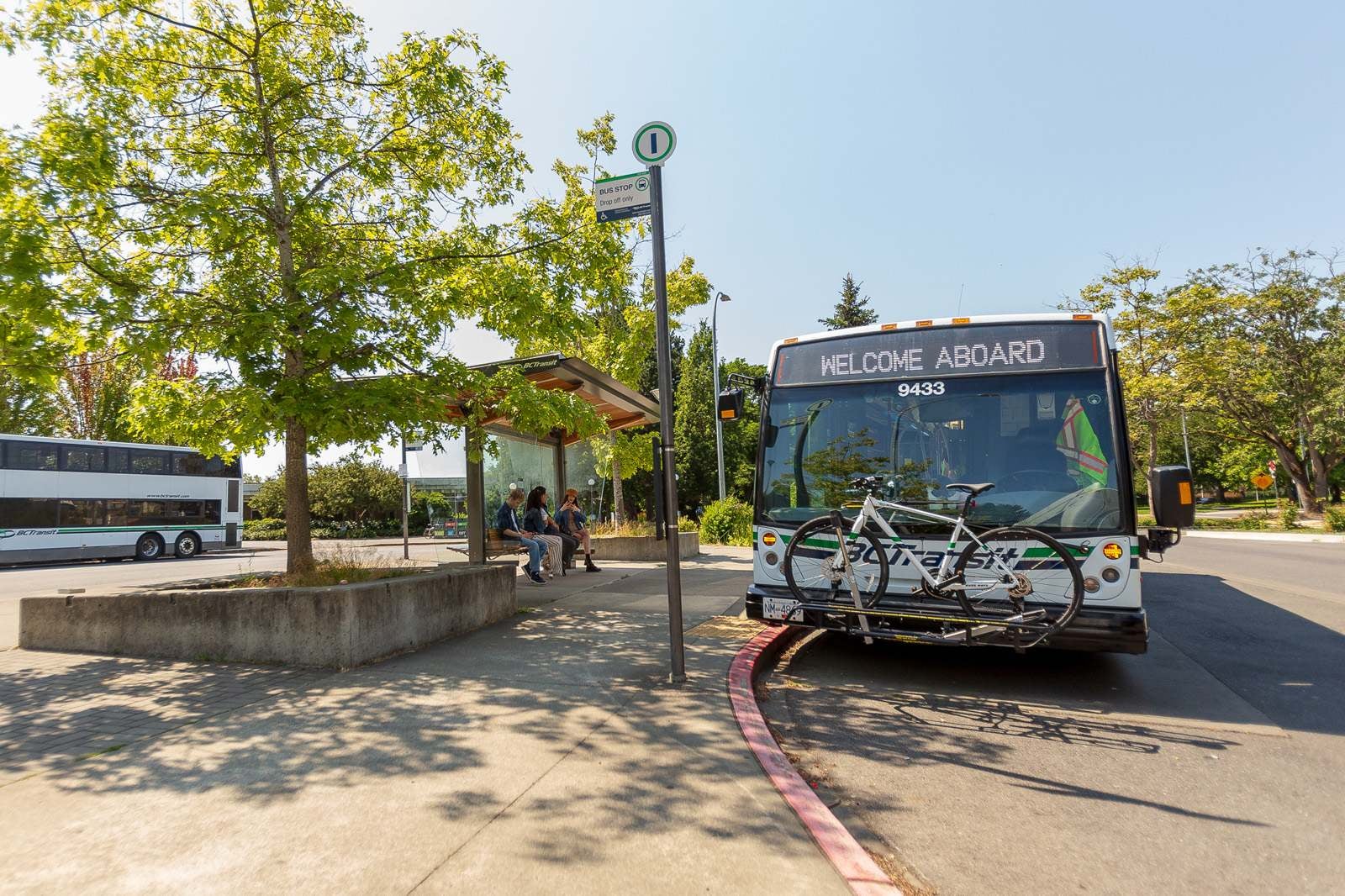 A BC Transit bus with a bike on the front rack is parked at a bus exchange. A double decker is parked in the background.