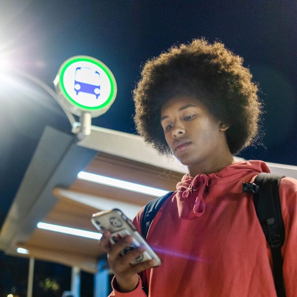 A rider checks their phone with an illuminated BC Transit sign and shelter behind.